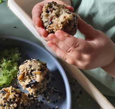 Child's hands holding sesame-coated rice ball over plate, mealtime mess-free with catch tray