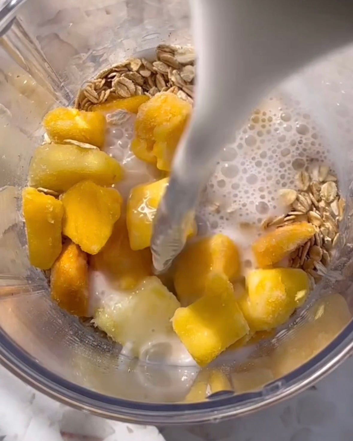 Milk pouring into blender with frozen mango chunks and oats for smoothie prep