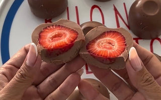 Hands holding chocolate-covered strawberry halves, revealing fresh red interior, with whole chocolates on plate