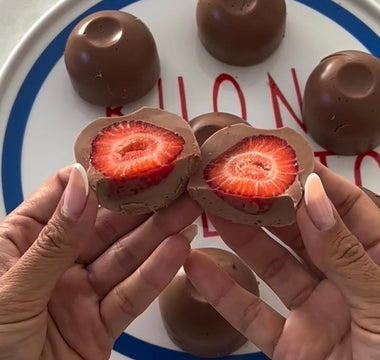 Hands holding chocolate-covered strawberry halves, revealing fresh red interior, with whole chocolates on plate