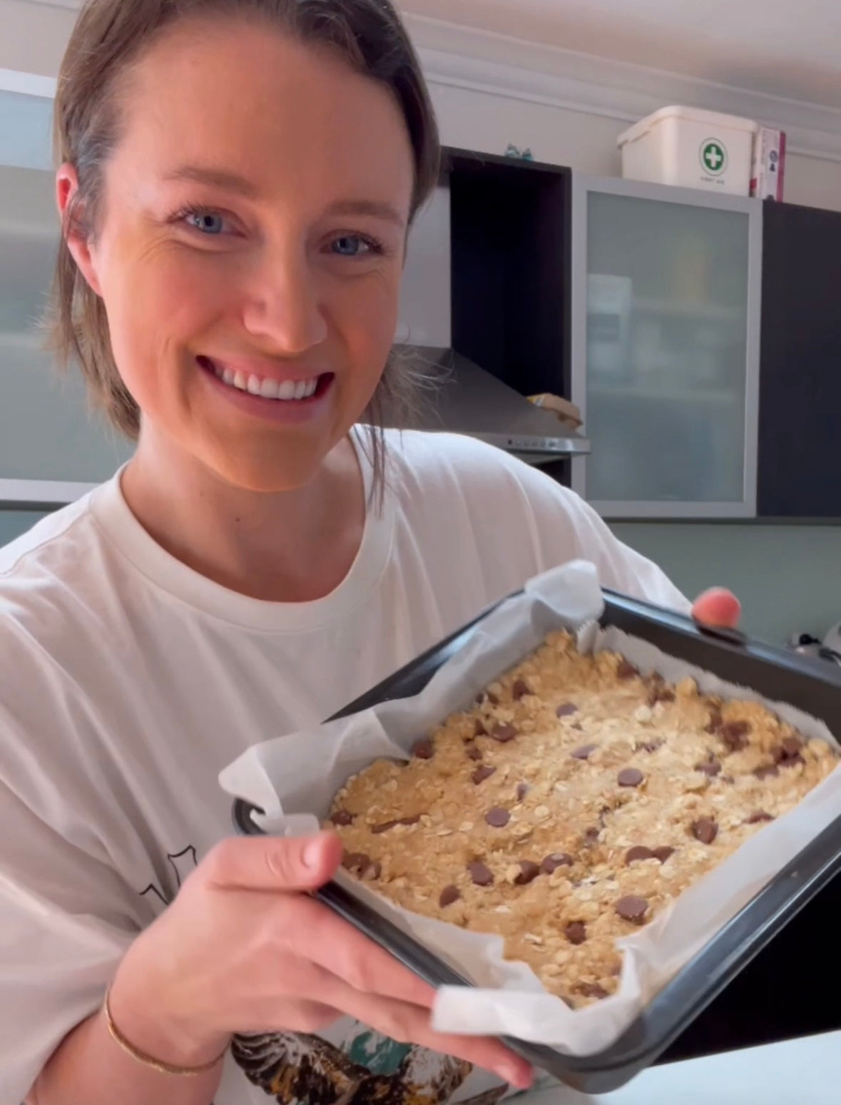Woman smiling while holding a baking tray with chocolate chip oat bars, ready to bake.