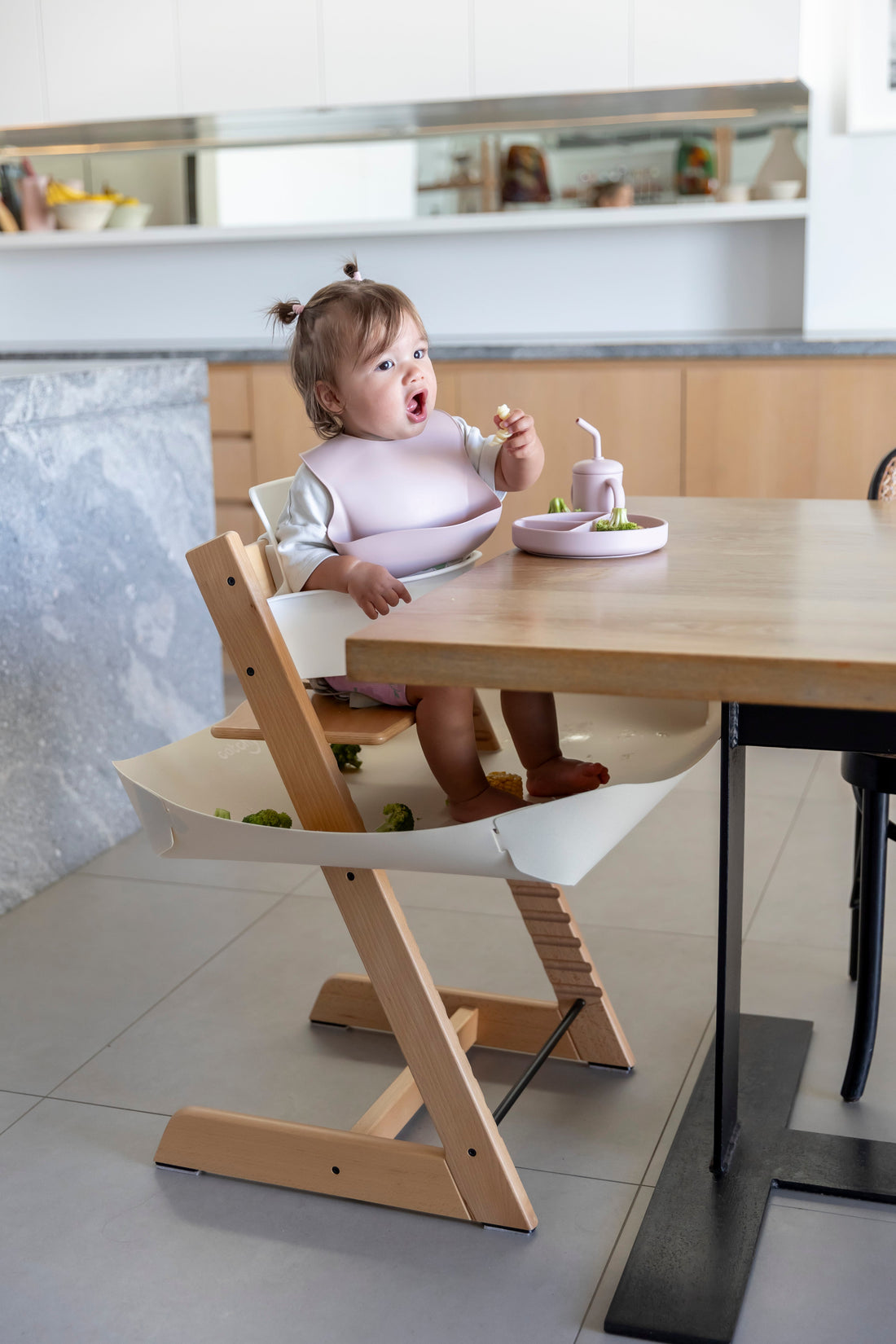 Baby eating with mess-free bib and food catcher under high chair, protecting floors during mealtime.