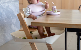 Baby eating with mess-free bib and food catcher under high chair, protecting floors during mealtime.